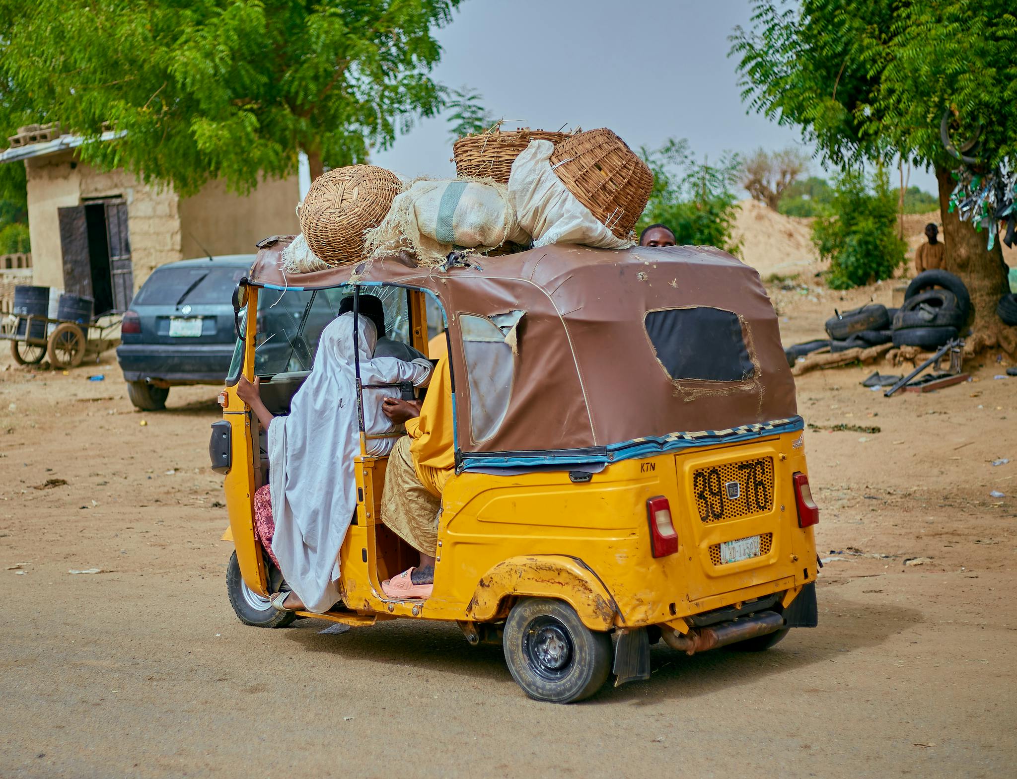 A yellow auto rickshaw loaded with baskets and passengers on a rural street.